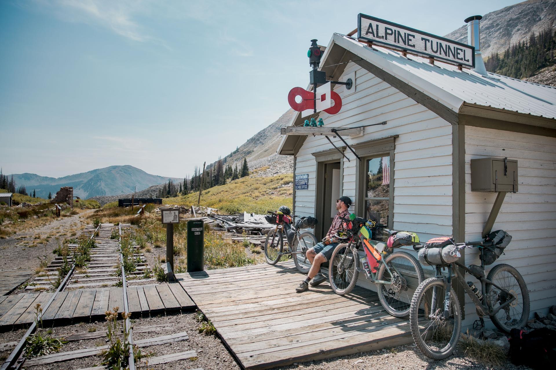 Person resting with bicycles on a wooden porch next to a small white building.