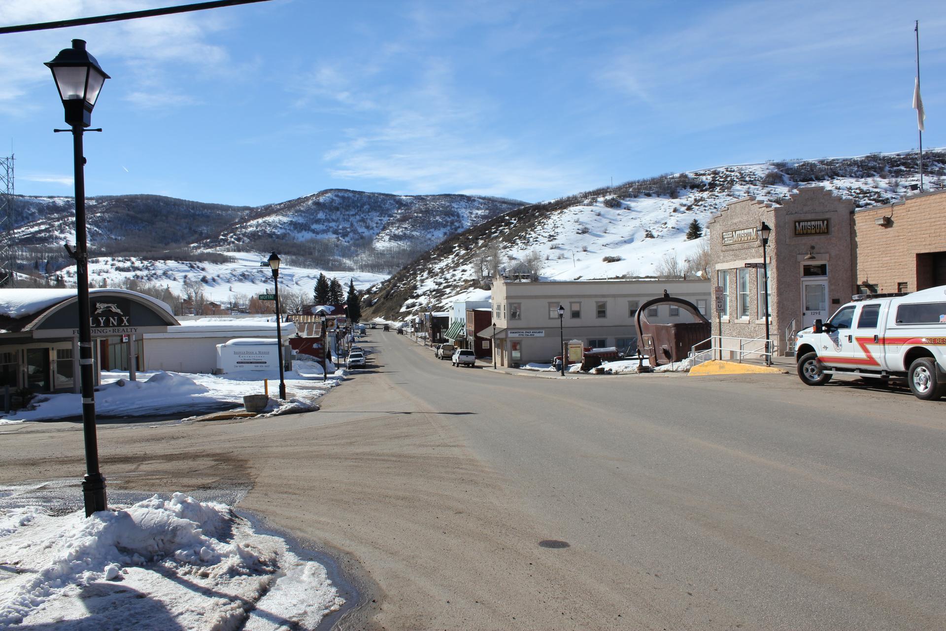 Sunny winter street scene with buildings, parked truck, and snow-covered mountains.