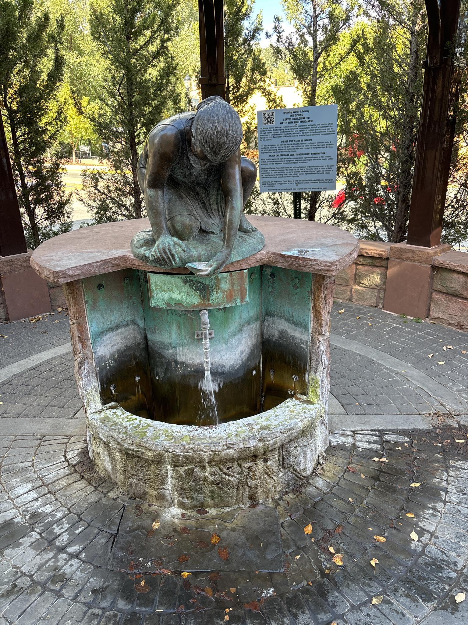 Bronze statue of a person kneeling over a stone fountain outdoors.