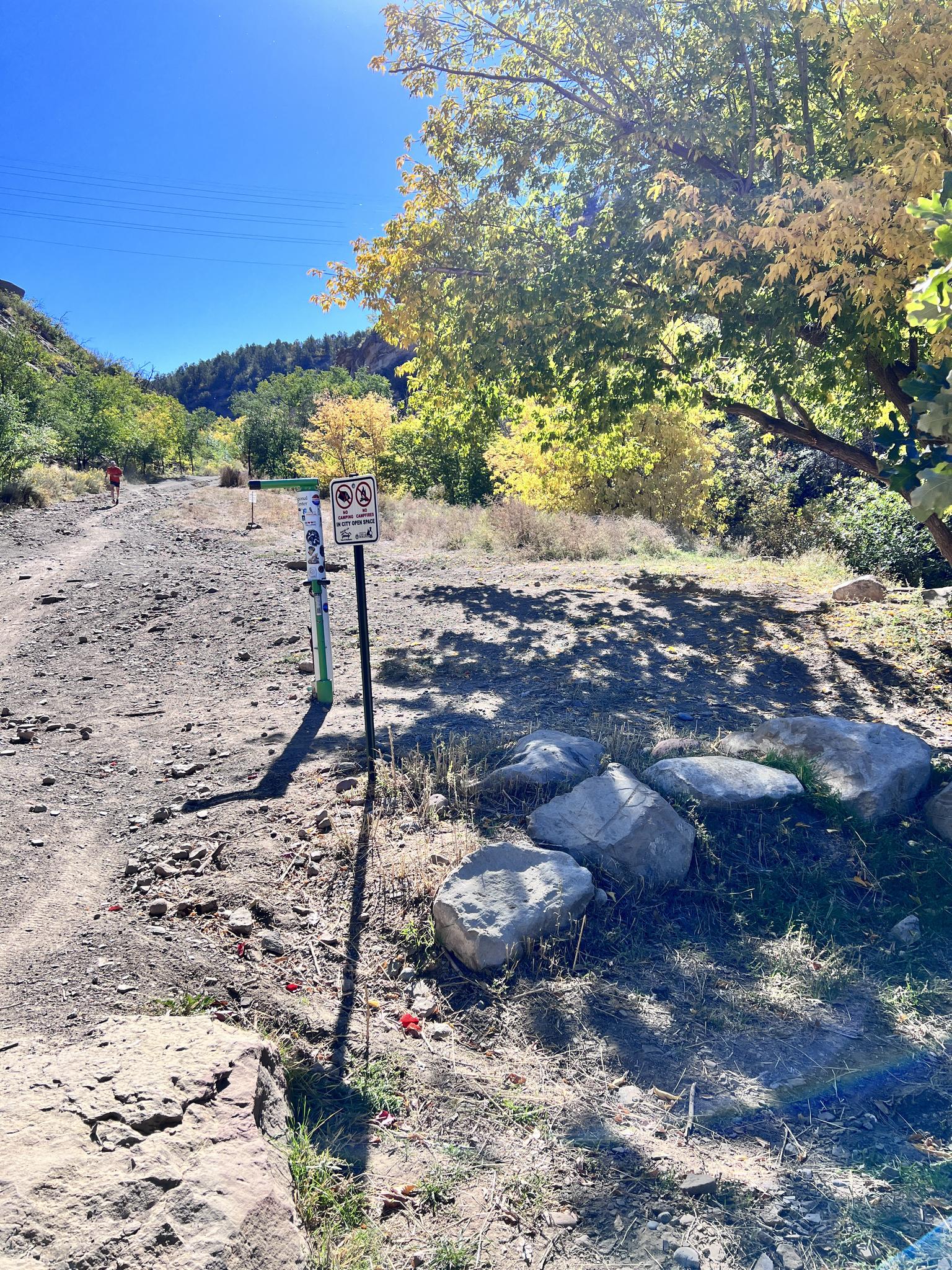 Dirt path with signs and large rocks under bright, leafy trees.