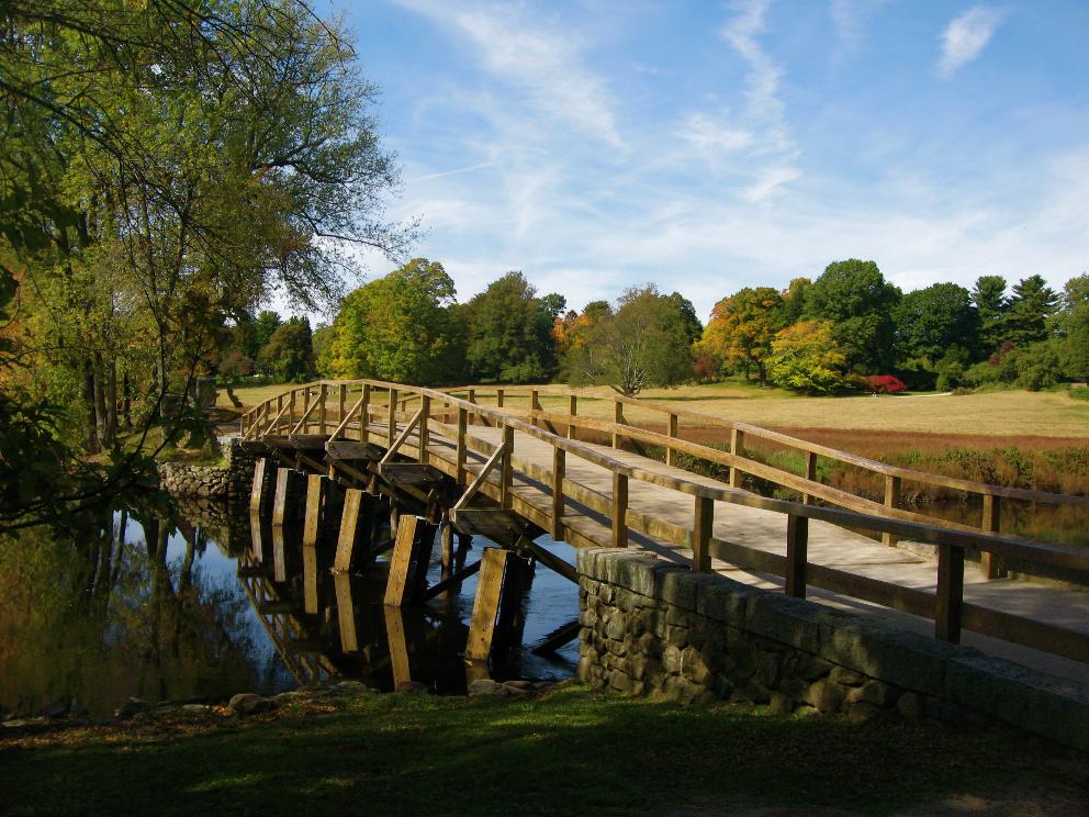 A rustic wooden bridge spans a calm river, surrounded by autumn trees.