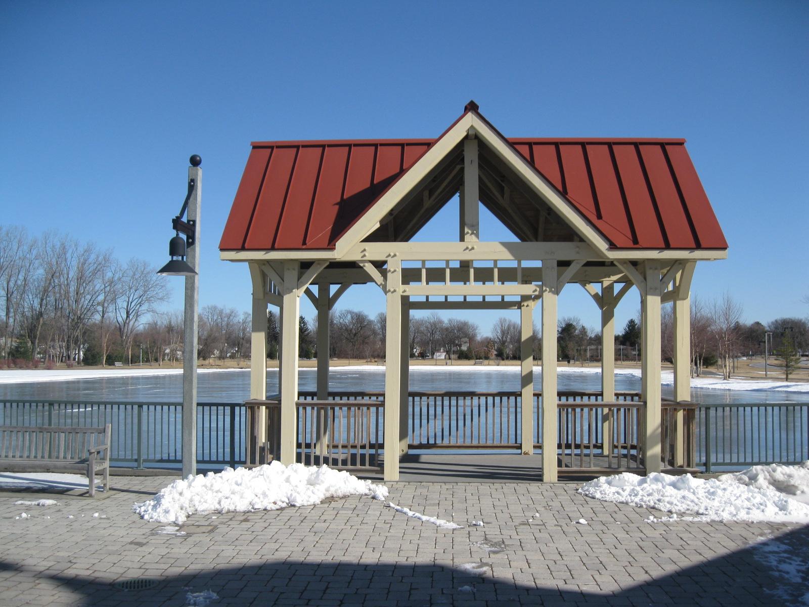 Sunny gazebo with red roof next to a frozen lake and snow.