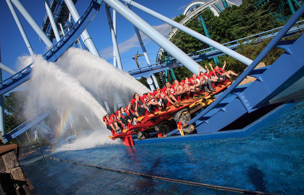 People on a roller coaster plunge into water, creating a large splash on a sunny day.