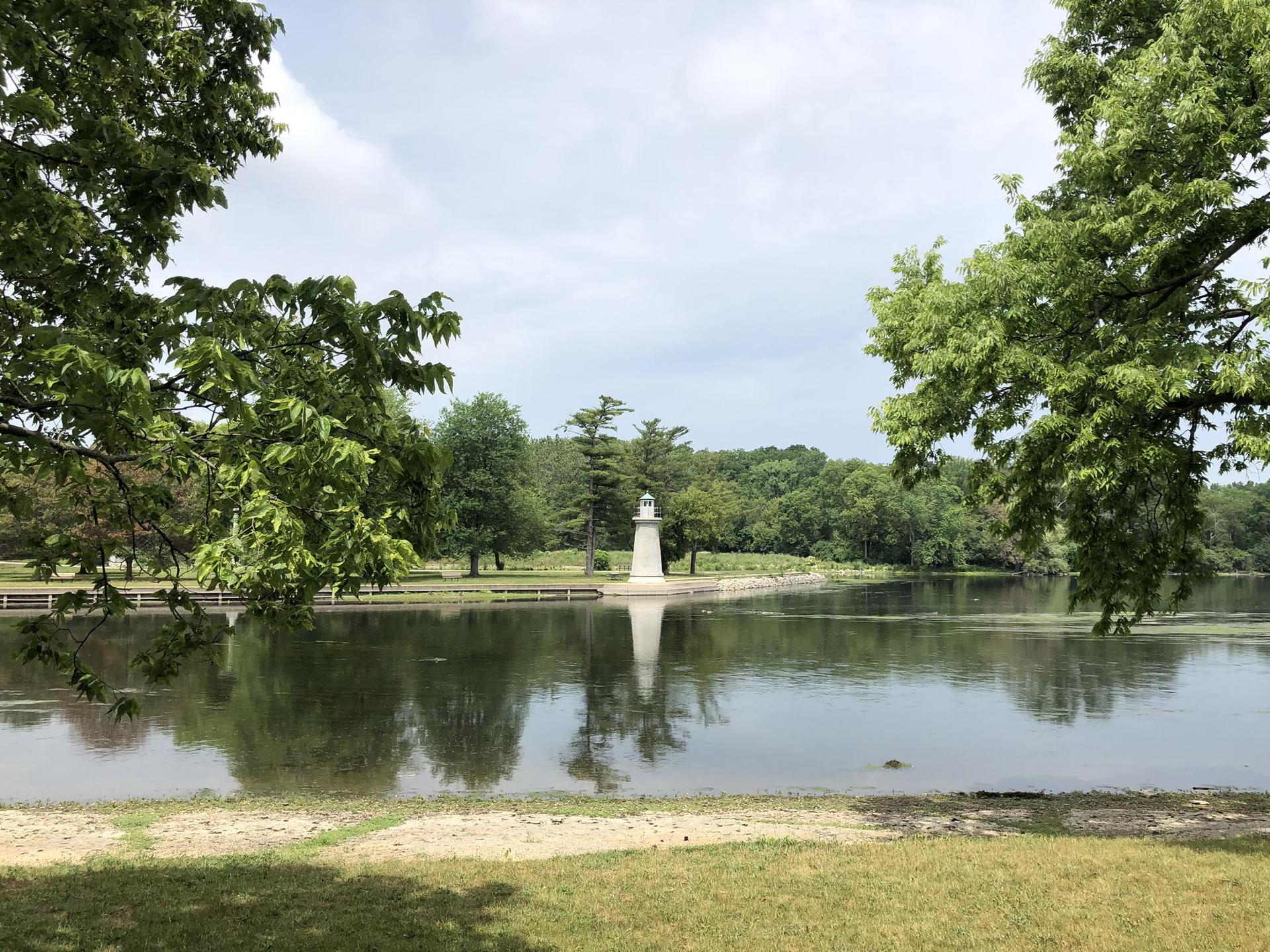 A white lighthouse stands in a calm lake, framed by green trees.