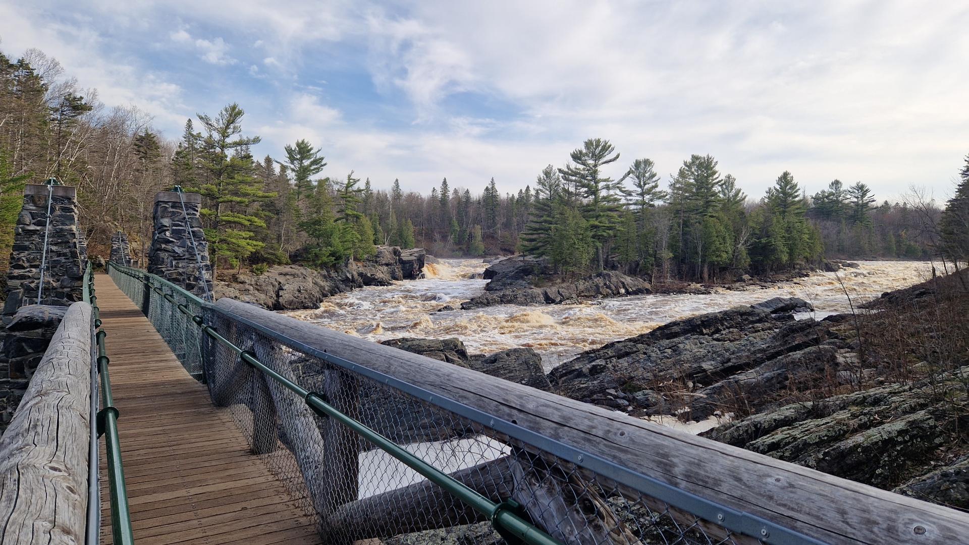 Wooden suspension bridge over a rocky river with rapids under a cloudy sky.