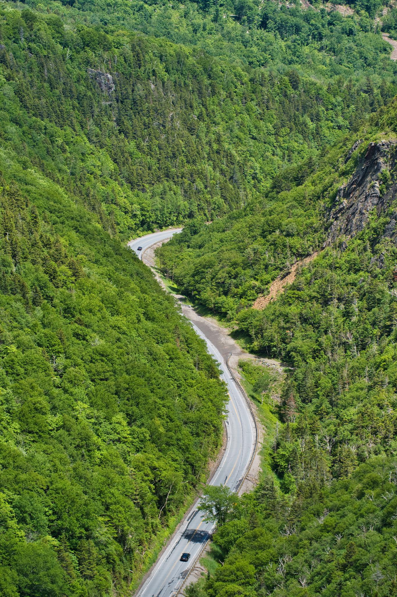 Winding road through a lush green valley with steep forested hillsides and cars.