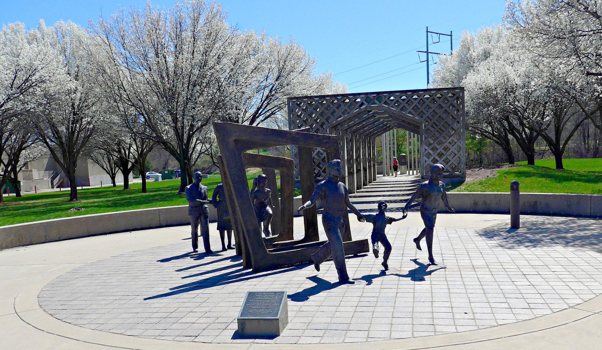 Bronze sculptures of people in a sunny park with white blossoming trees.