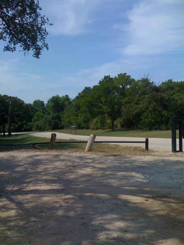 Sunny dirt path and barrier leading to a paved road, surrounded by green trees.