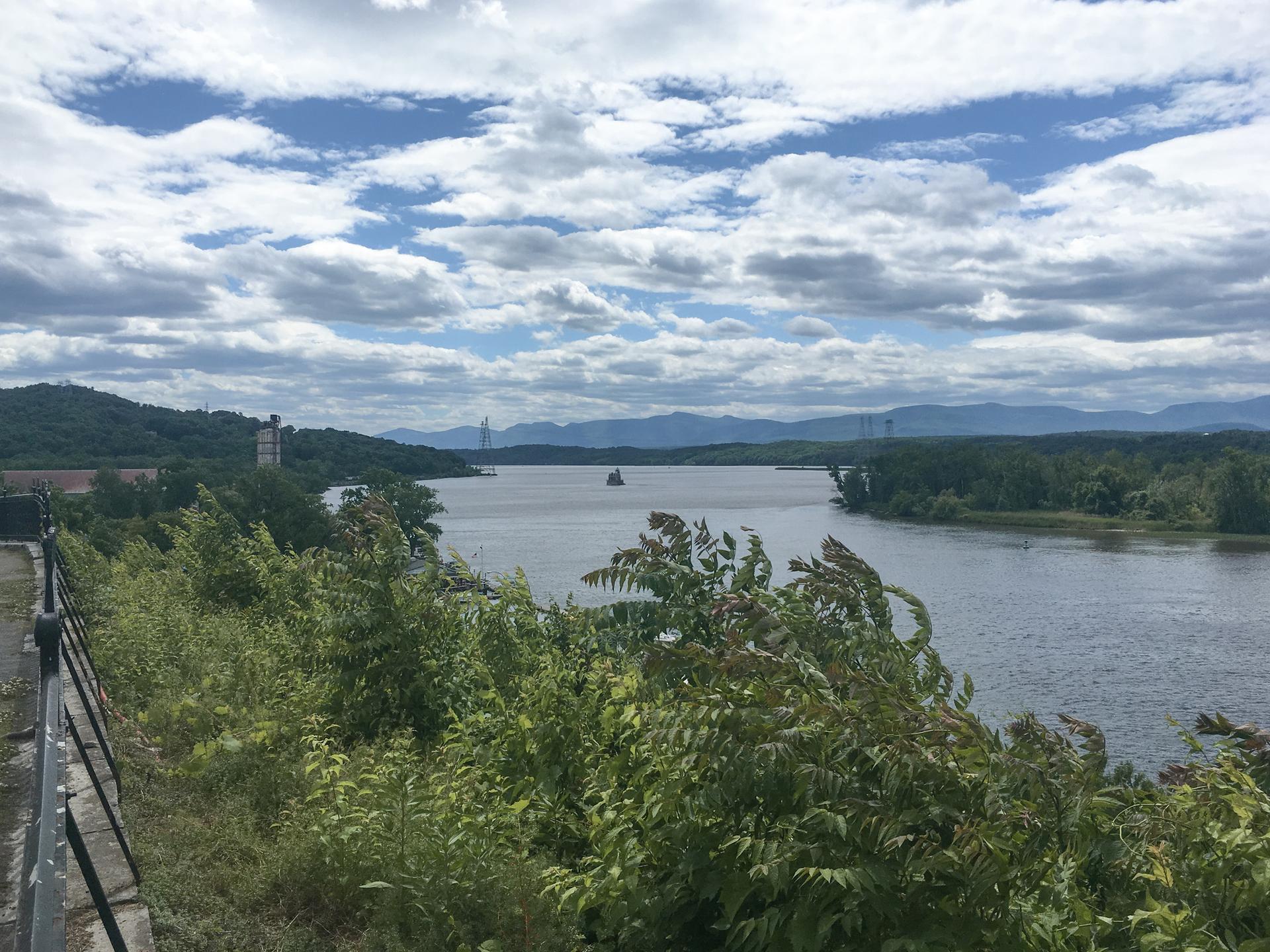 A wide river with distant mountains under a cloudy sky, seen through foliage.