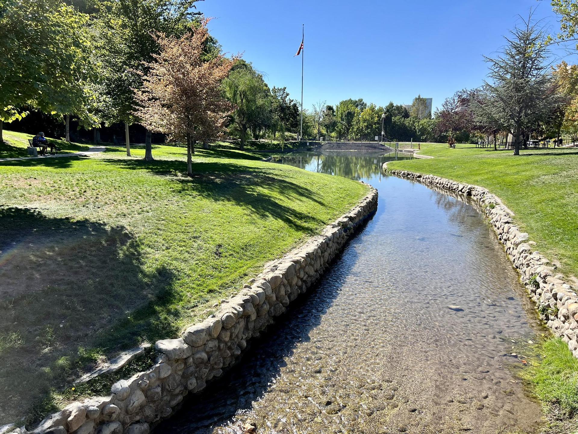 A clear stream flows through a sunny green park with trees and a bridge.