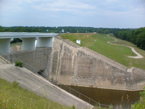 Large concrete dam with a bridge, grassy slopes, and dark water under overcast sky.