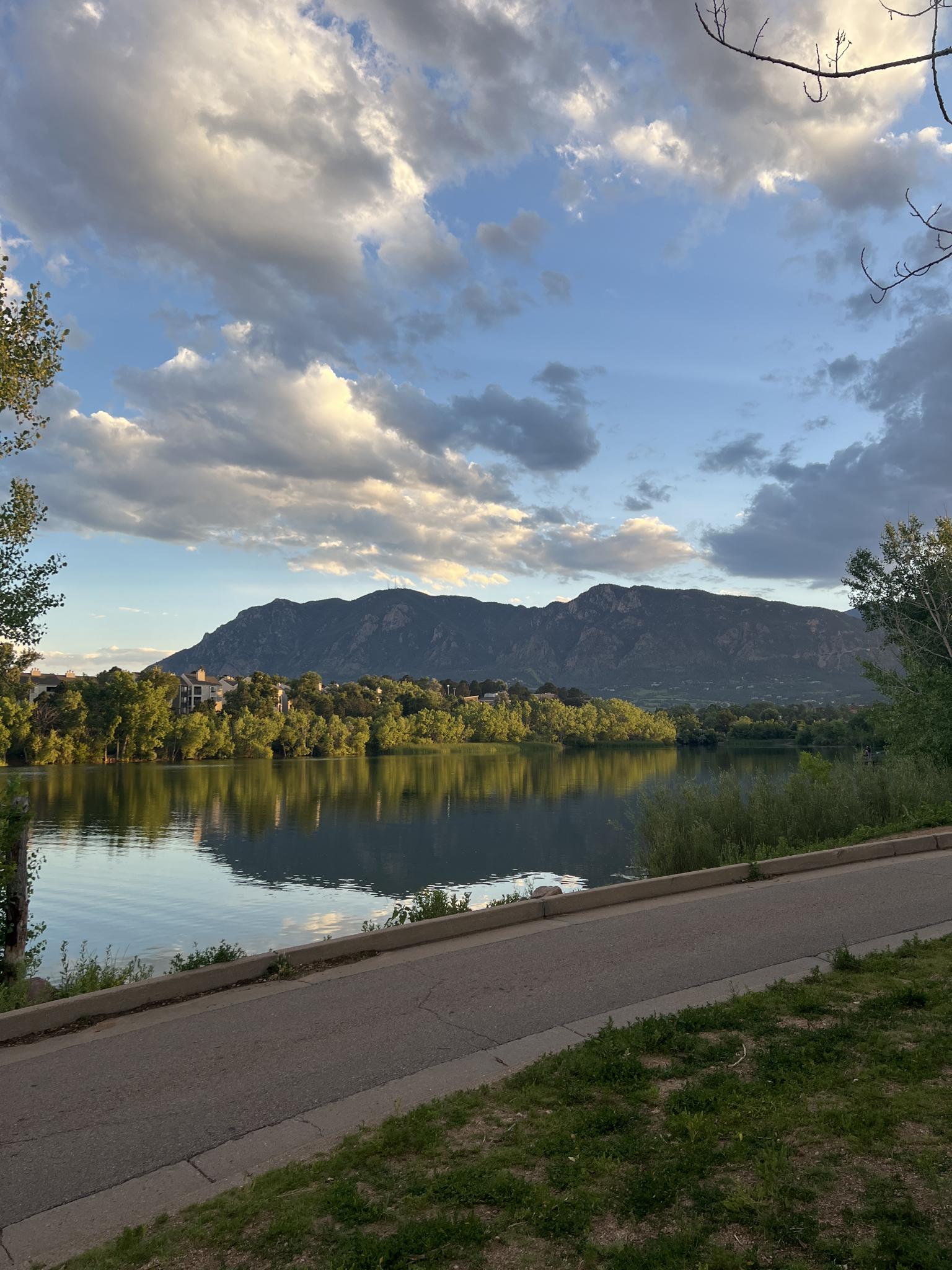 Calm lake reflecting mountains and green trees, with a paved path.