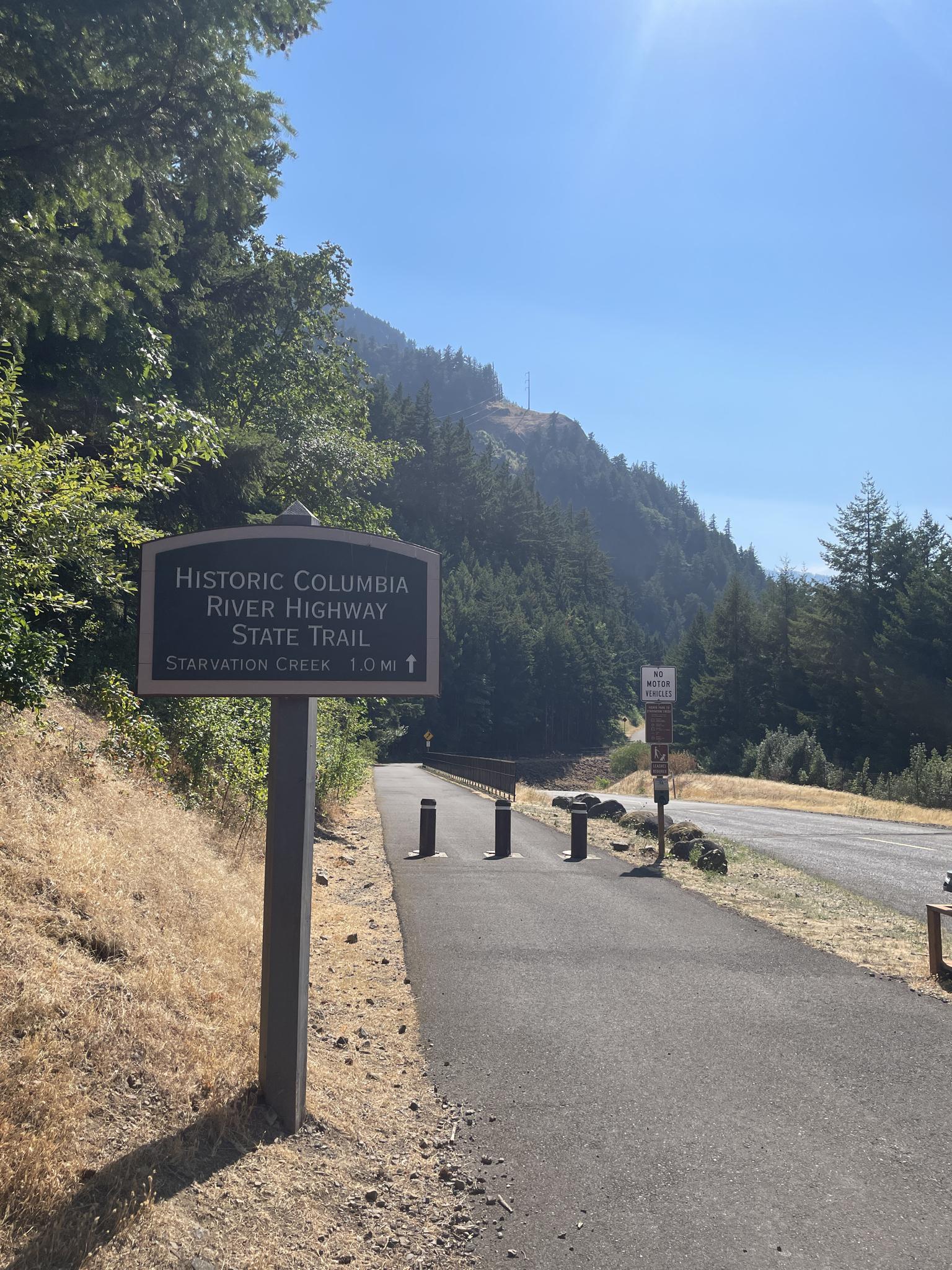 A trail sign stands beside a paved path leading into sunny, forested mountains.