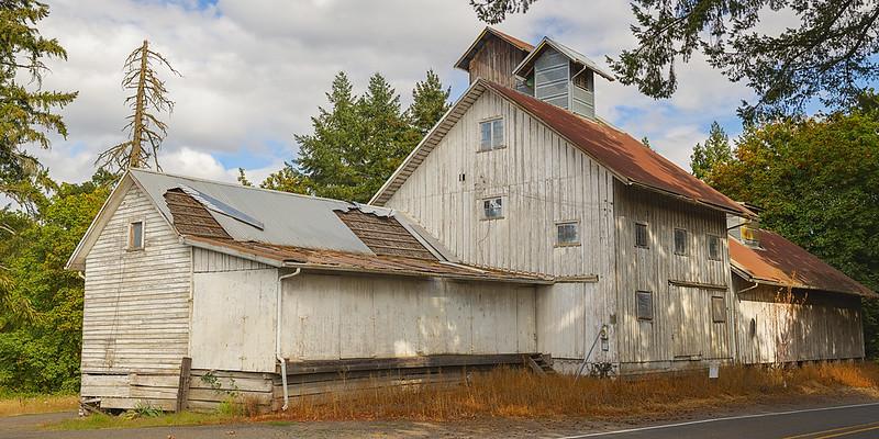 An old white barn with a red roof stands among trees under an overcast sky.