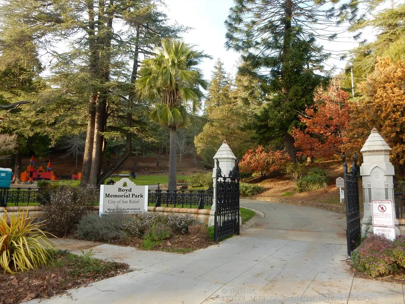 Stone pillars mark a park entrance with tall trees and green foliage.