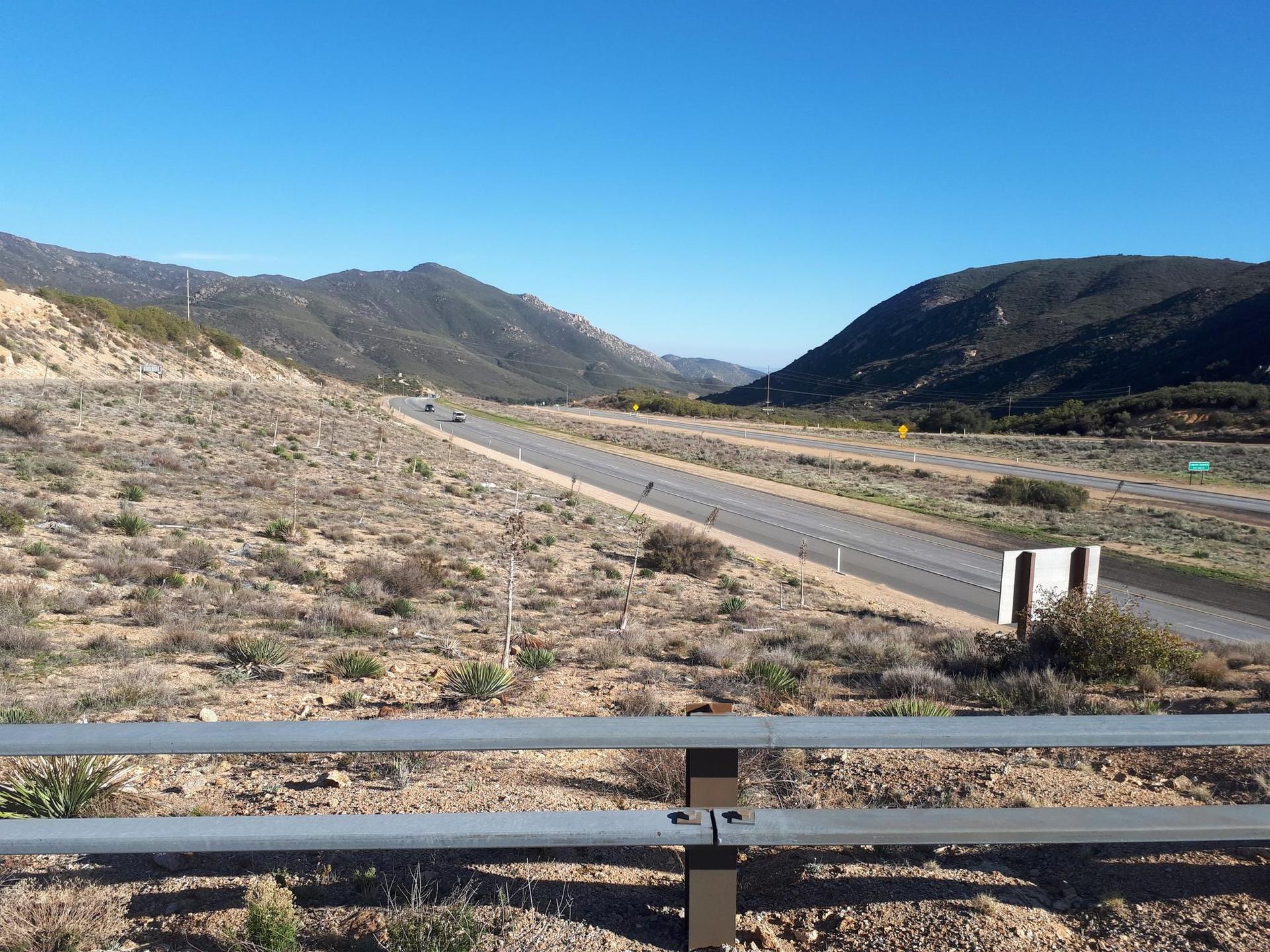 Highway cutting through dry, brushy hills under a clear blue sky.