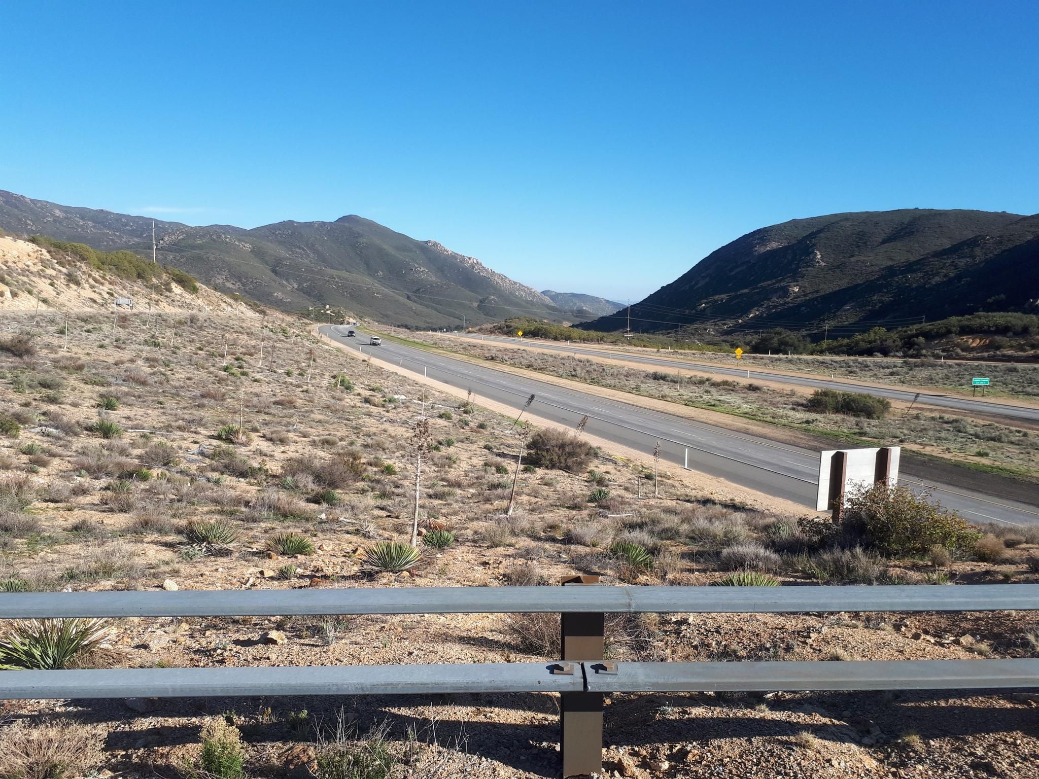 Highway cutting through dry, brushy hills under a clear blue sky. - Wooded Hill Loop