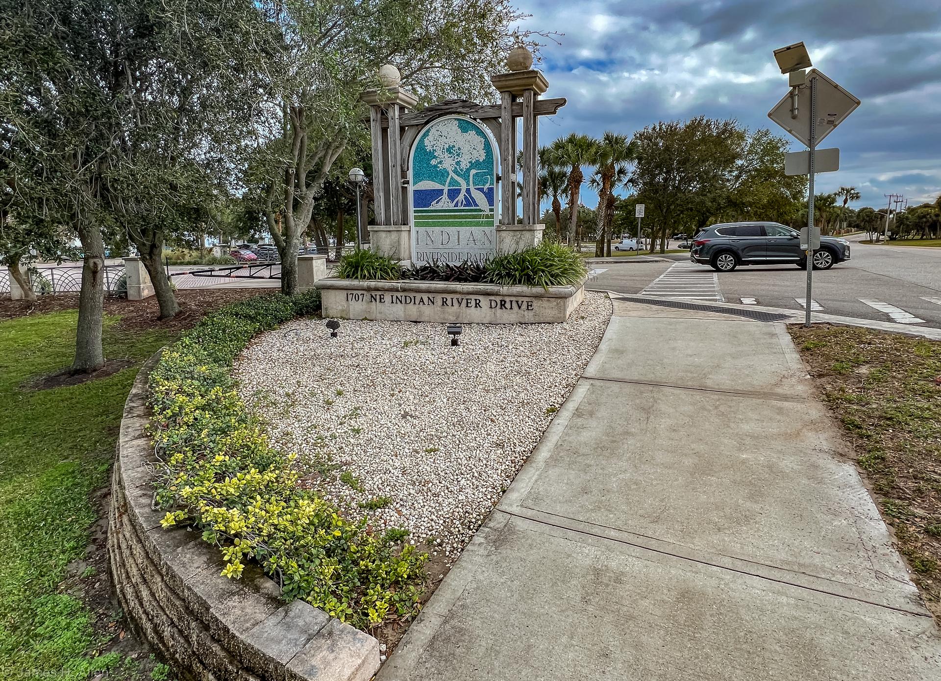 Stone monument with tree logo, gravel, and road under an overcast sky.