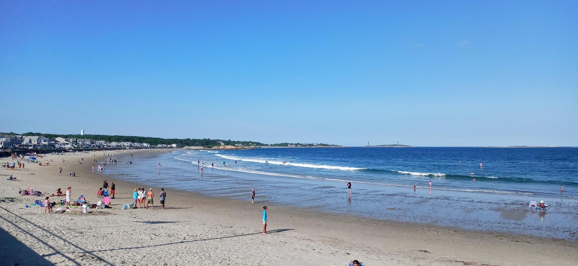 Busy sandy beach with many people enjoying ocean waves under a clear sky.