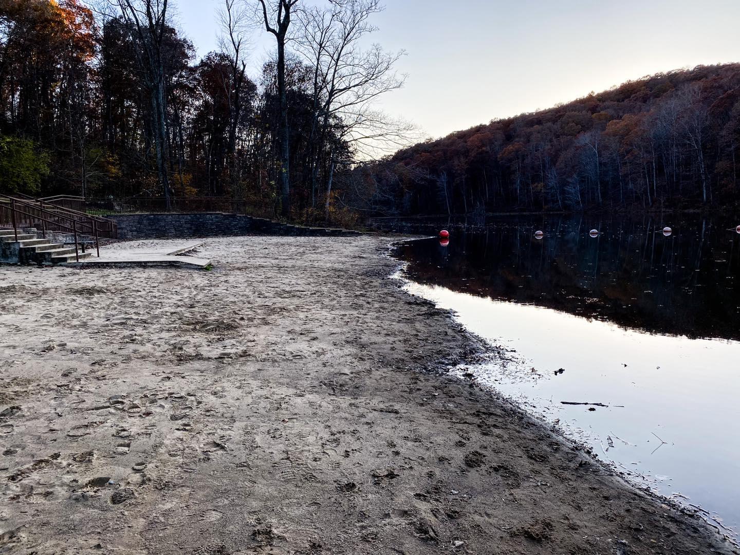 A sandy beach next to a dark lake with forested hills under an overcast sky.