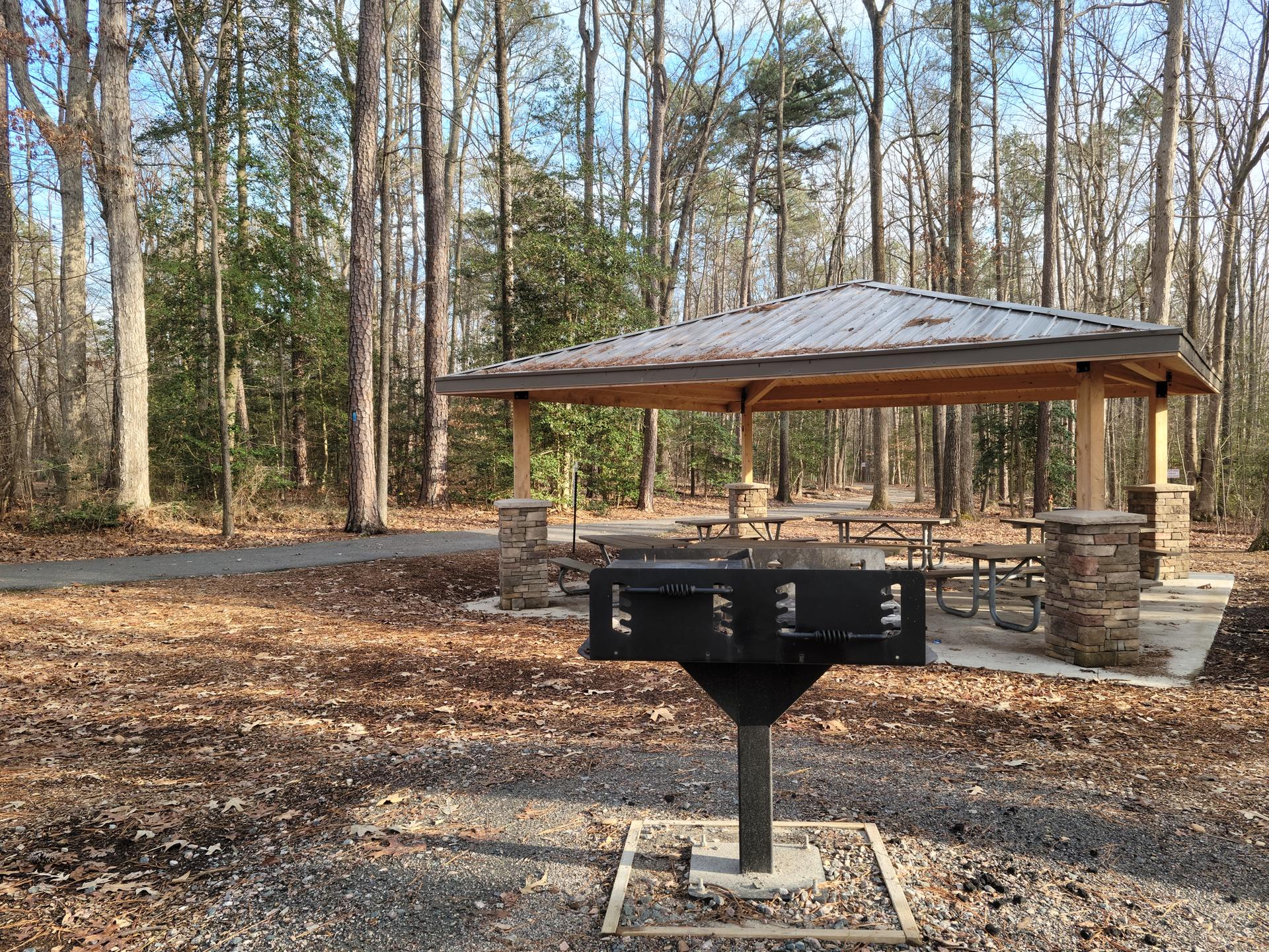 A black grill in front of a covered picnic shelter in a sunny wooded area.