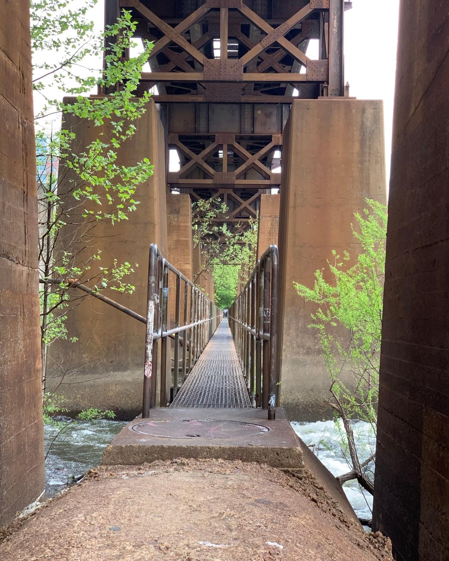 Narrow metal walkway under a large, rusty bridge structure over water.
