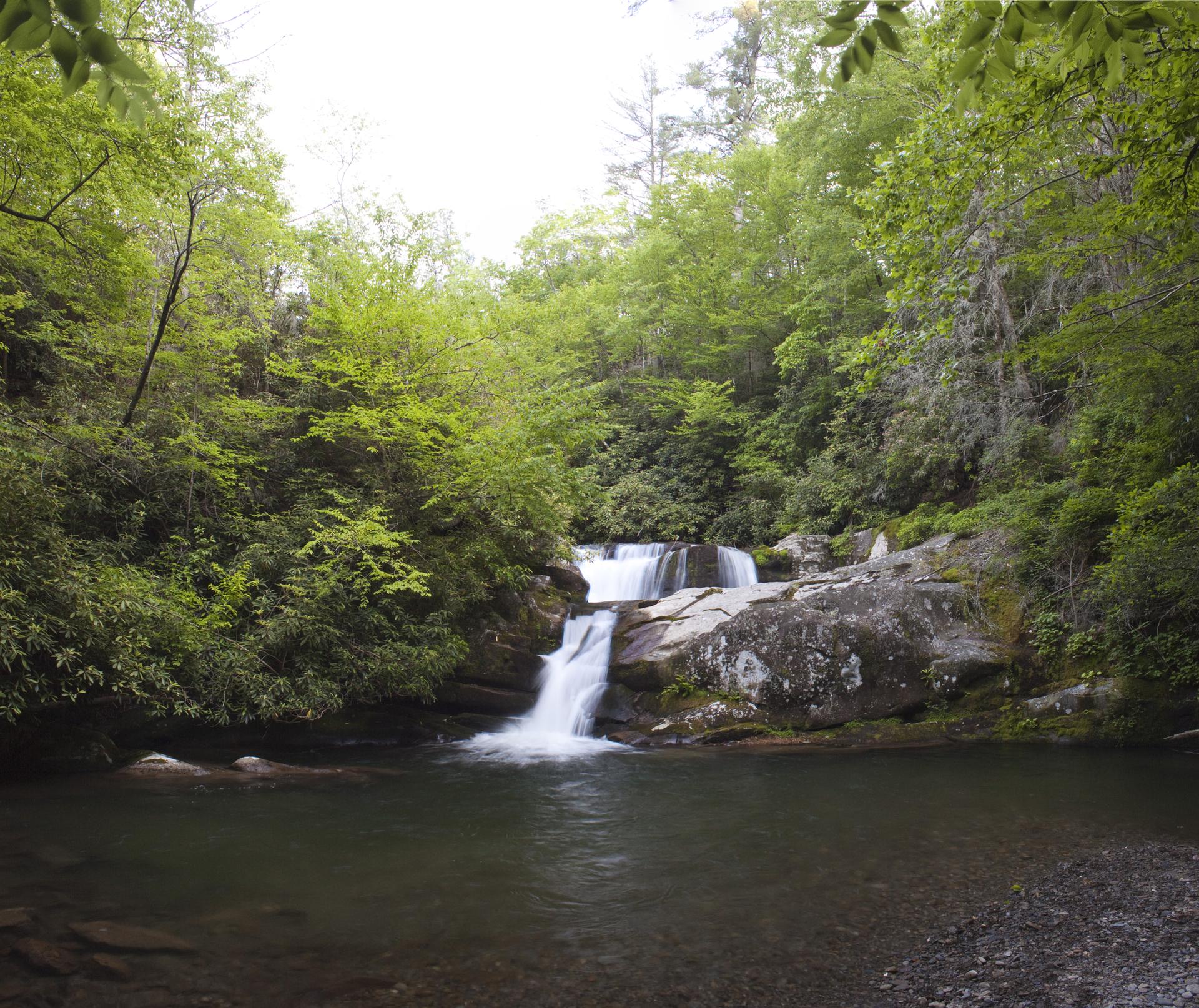 A multi-tiered waterfall cascades into a dark pool surrounded by lush green forest.