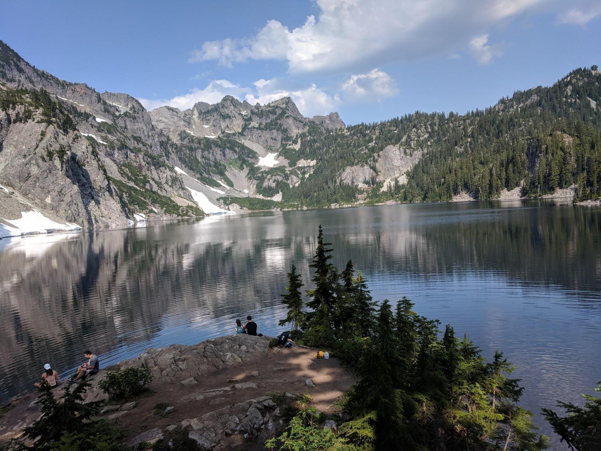 People relax by a calm mountain lake reflecting rocky peaks under a bright sky.