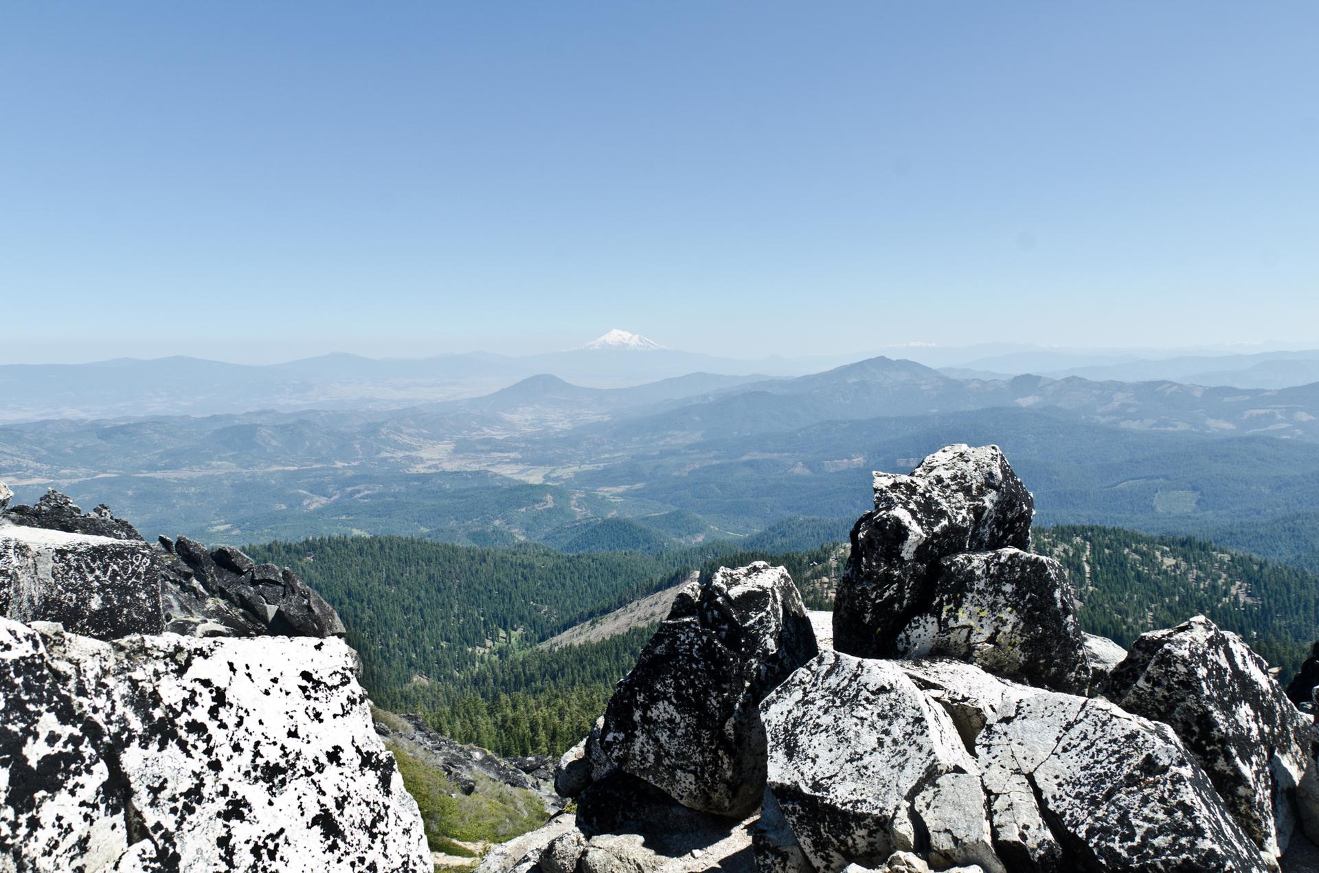 Mountain peak with large rocks, overlooking a vast forested landscape under clear skies.