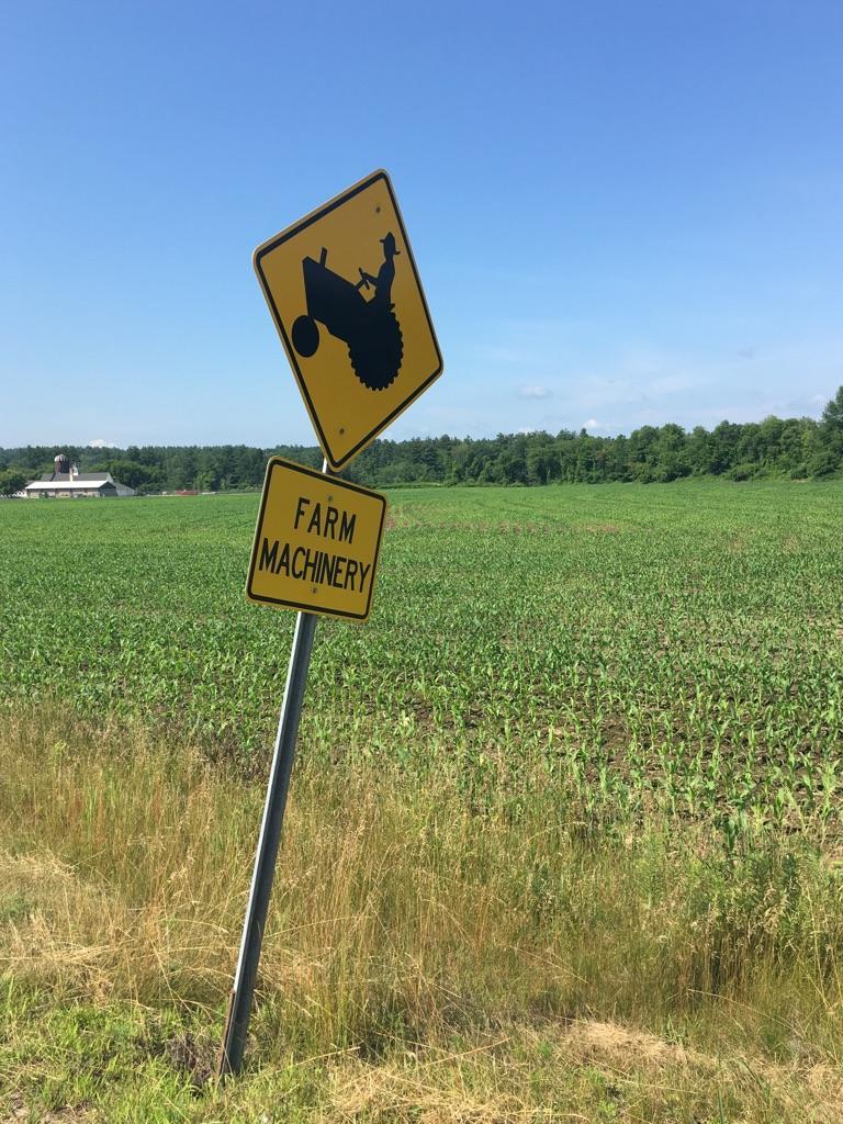 A yellow farm machinery warning sign stands beside a green crop field.