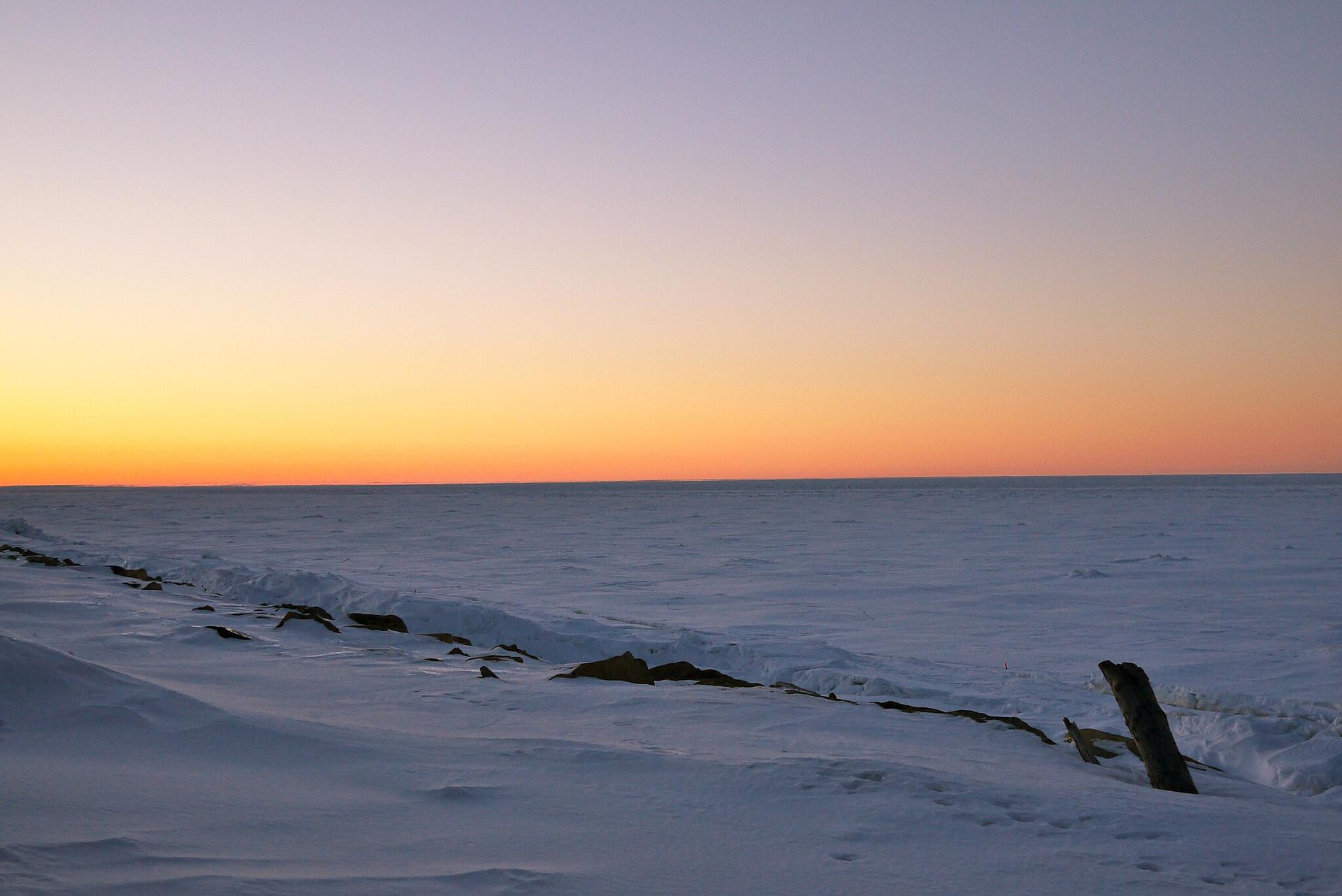 Vast snowy and icy landscape under a colorful sunset sky, fading to dark.