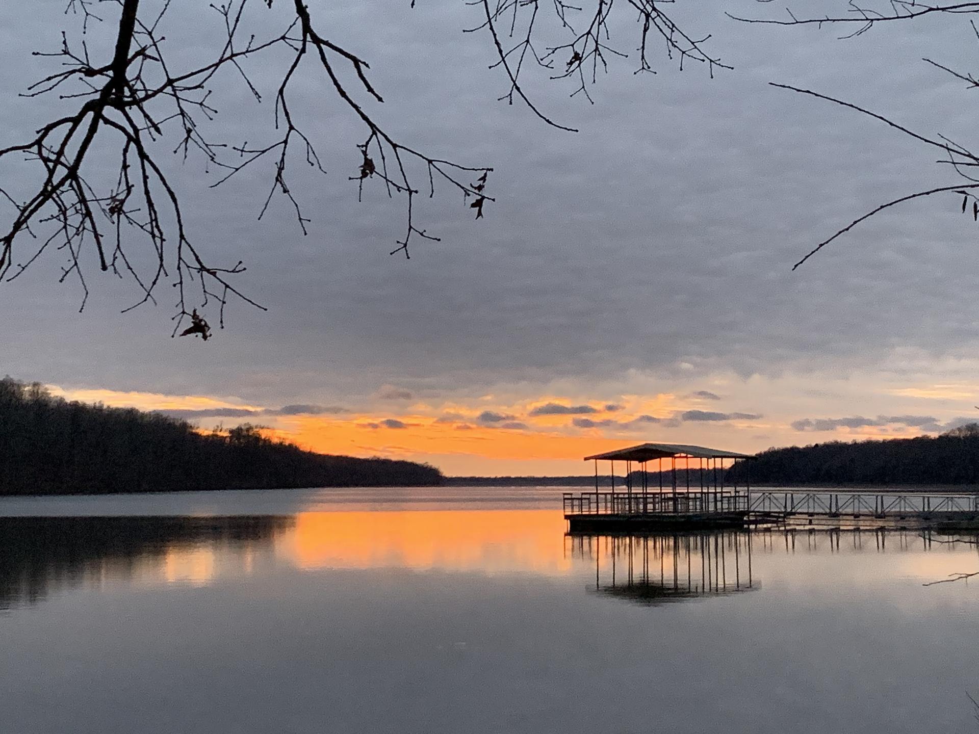 Orange sunset over a calm lake with a covered dock and silhouetted trees.