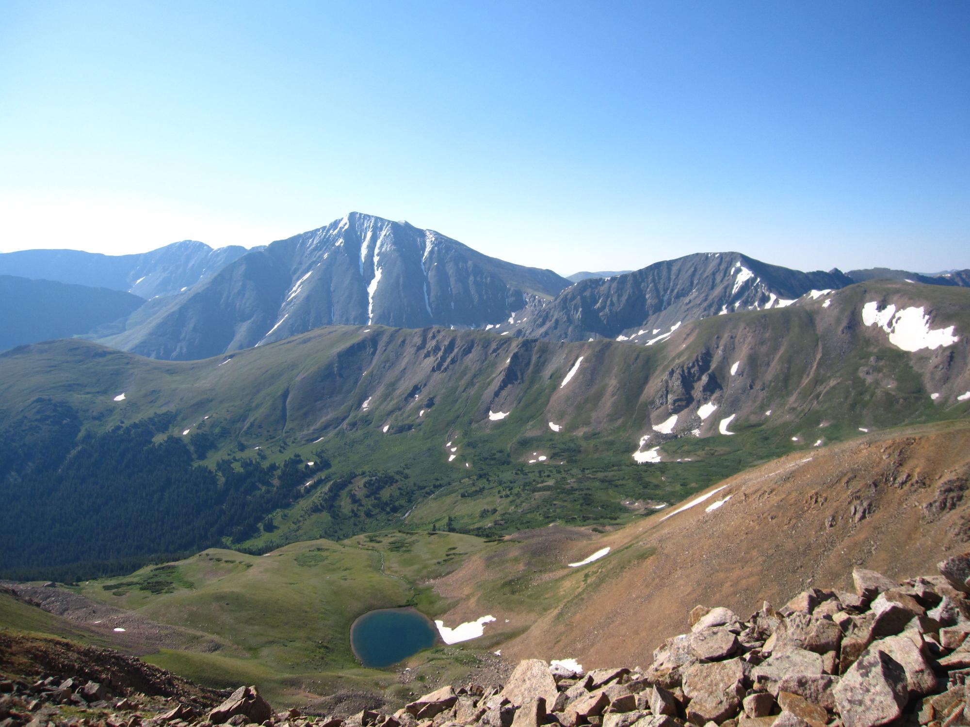 Bright mountain landscape with snow-dusted peaks, green valleys, and a small lake.