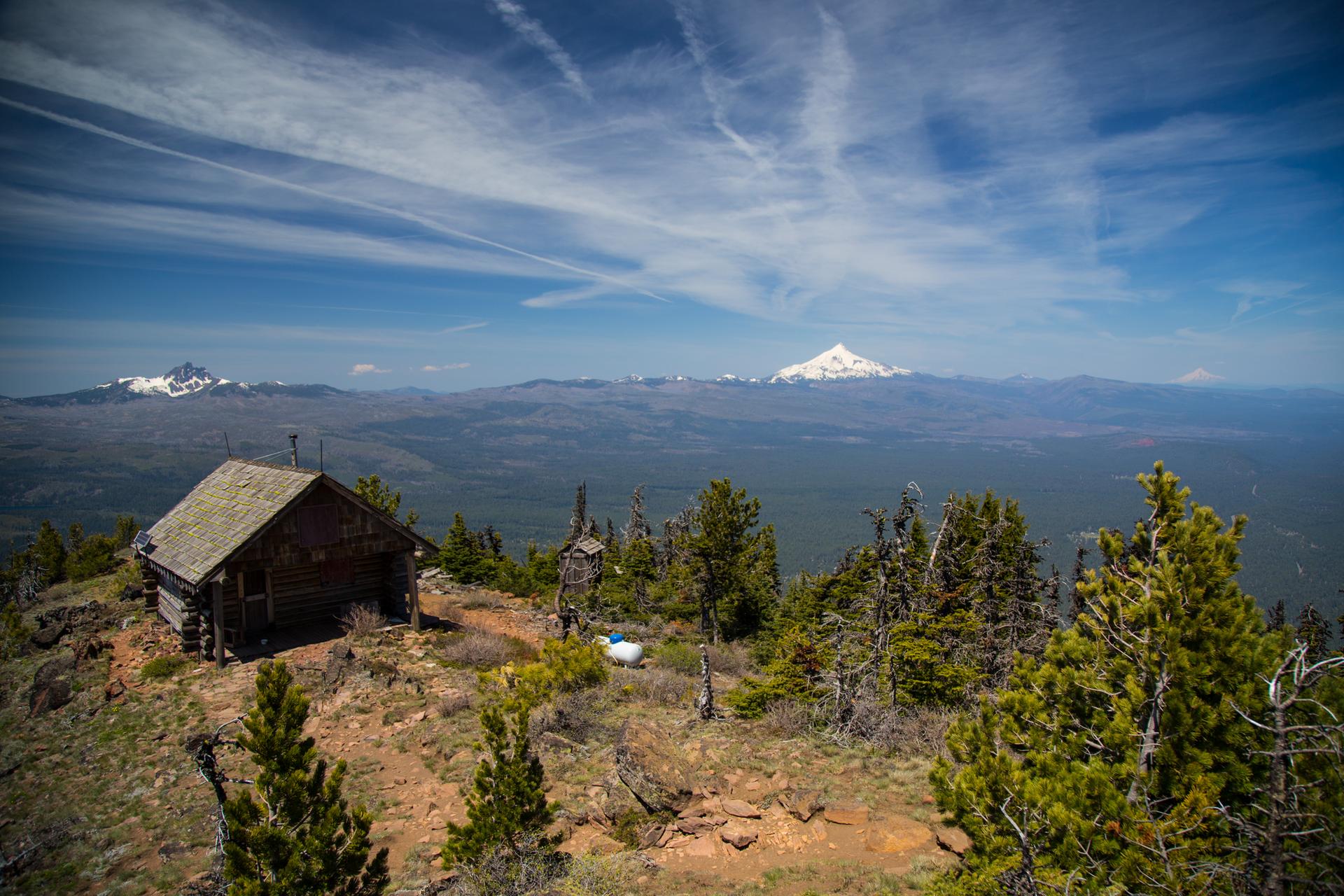 A rustic cabin sits on a mountain peak with distant snow-capped mountains.