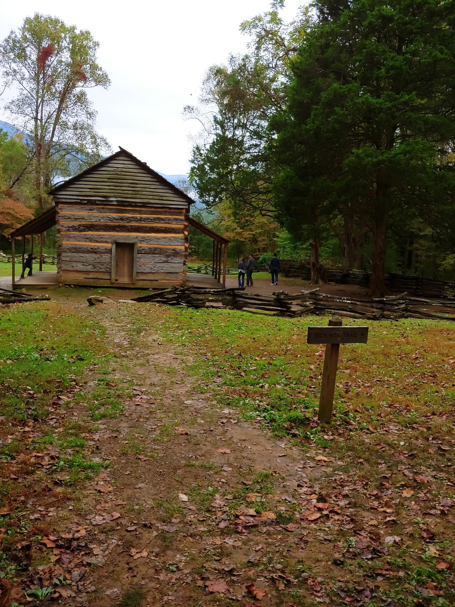 Historic log cabin with people exploring a wooded area on an overcast day.