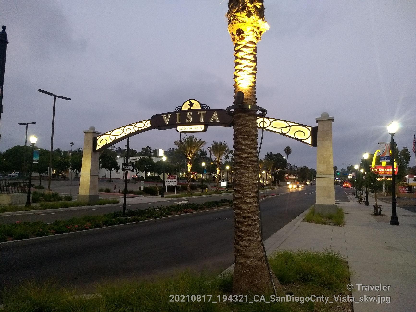 Illuminated archway over a street at dusk, with palm trees and lights.