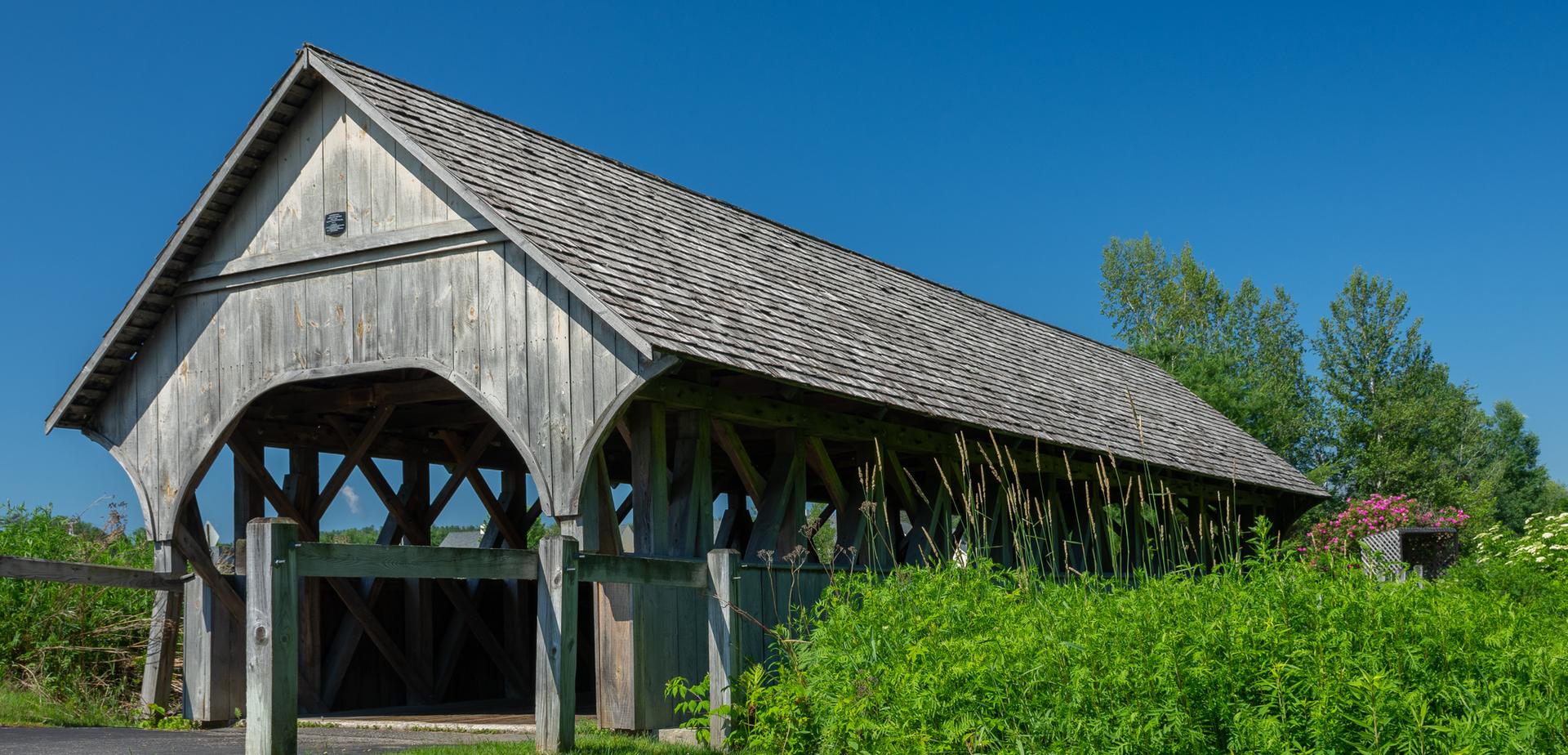 A rustic wooden covered bridge under a clear blue sky, surrounded by green plants.