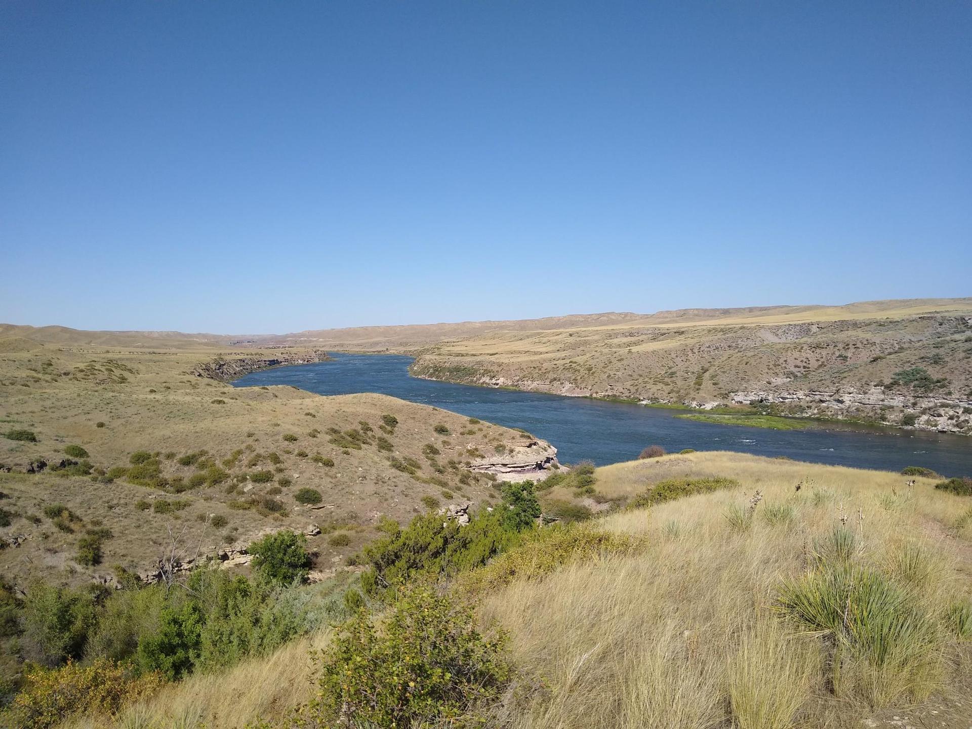 River winds through dry, grassy hills under a clear blue sky.