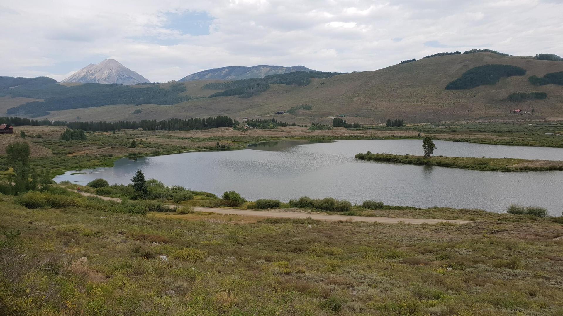 Calm lake surrounded by grassy hills and distant mountains under cloudy sky.