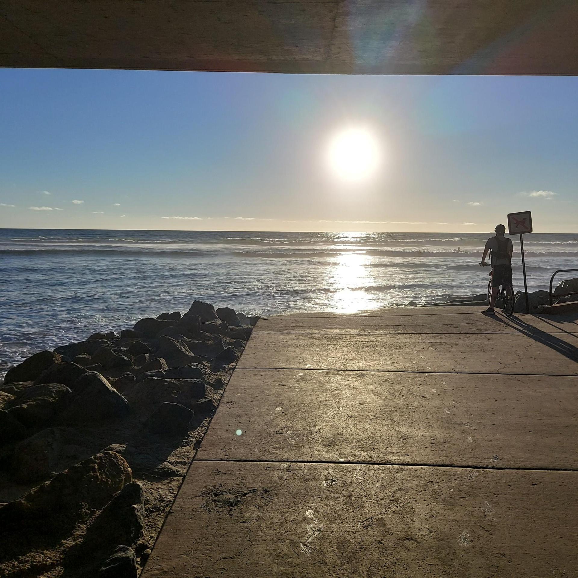 Person with bike on concrete path watching bright sunset over ocean waves.