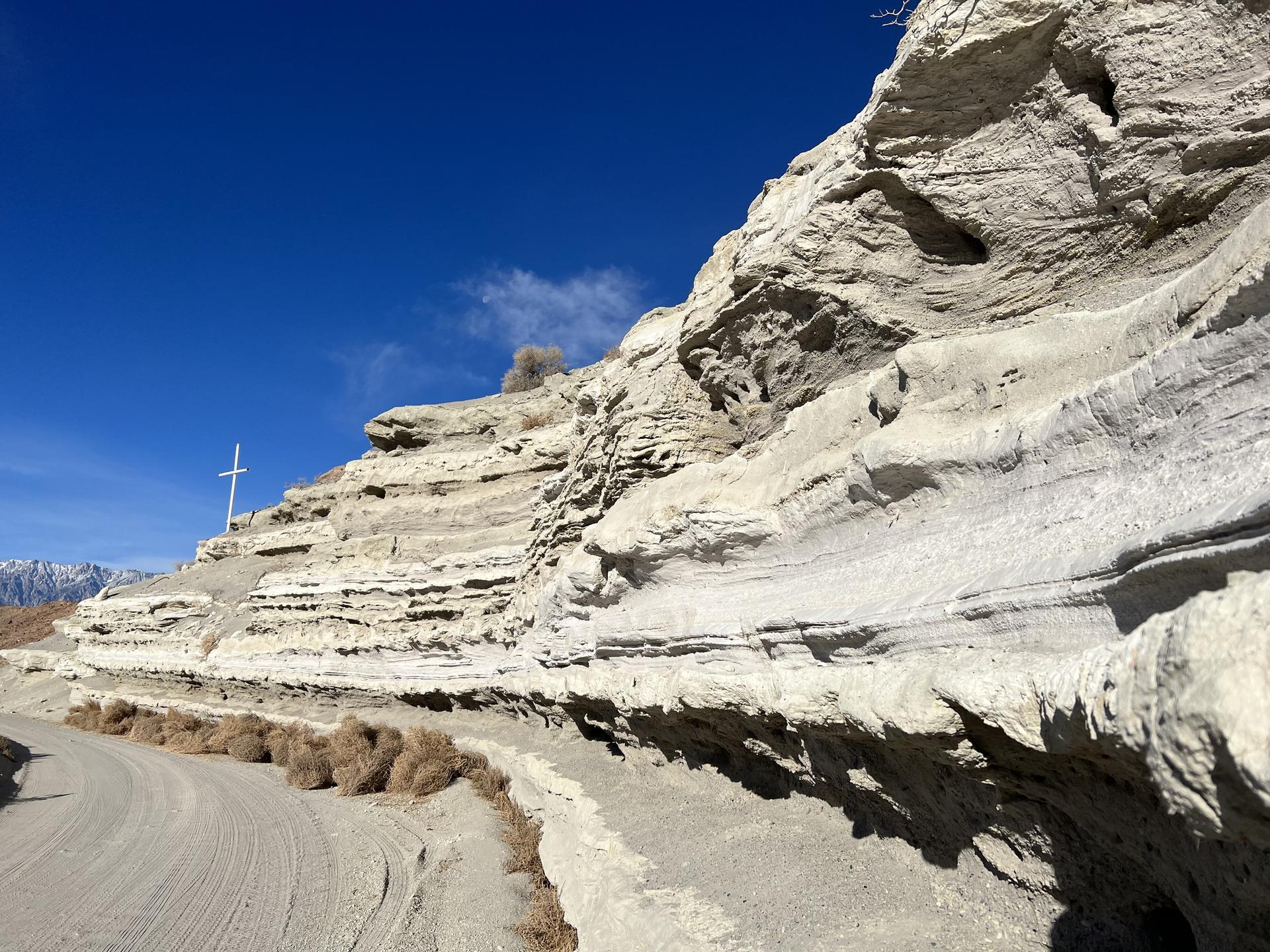 A white cross stands on layered rock formations beside a dirt road.