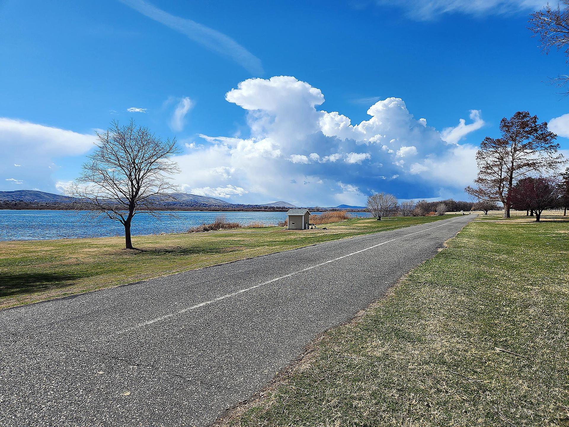 A paved road runs beside a lake with bare trees and dramatic white clouds.