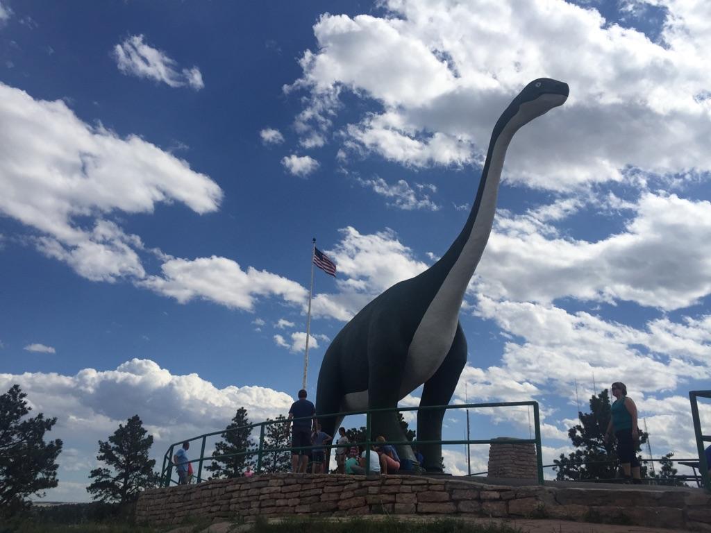 People gather around a large dinosaur statue under a bright, cloudy sky.