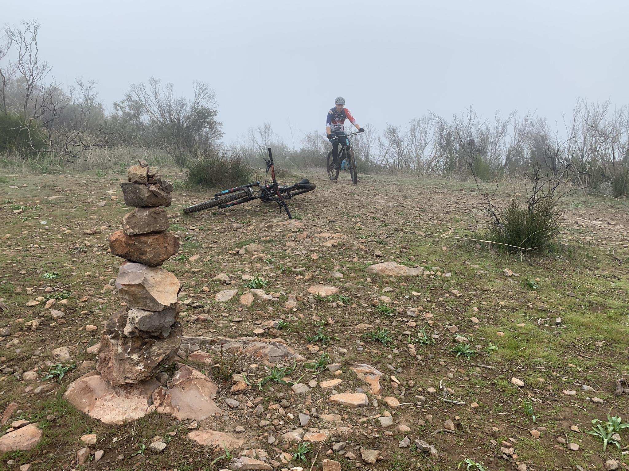 Mountain biker and bike near a rock cairn on a foggy, rocky trail.