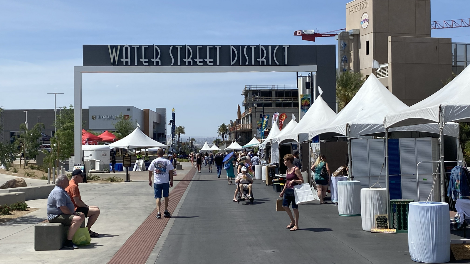Busy outdoor market with white tents, people walking, and a large sign.