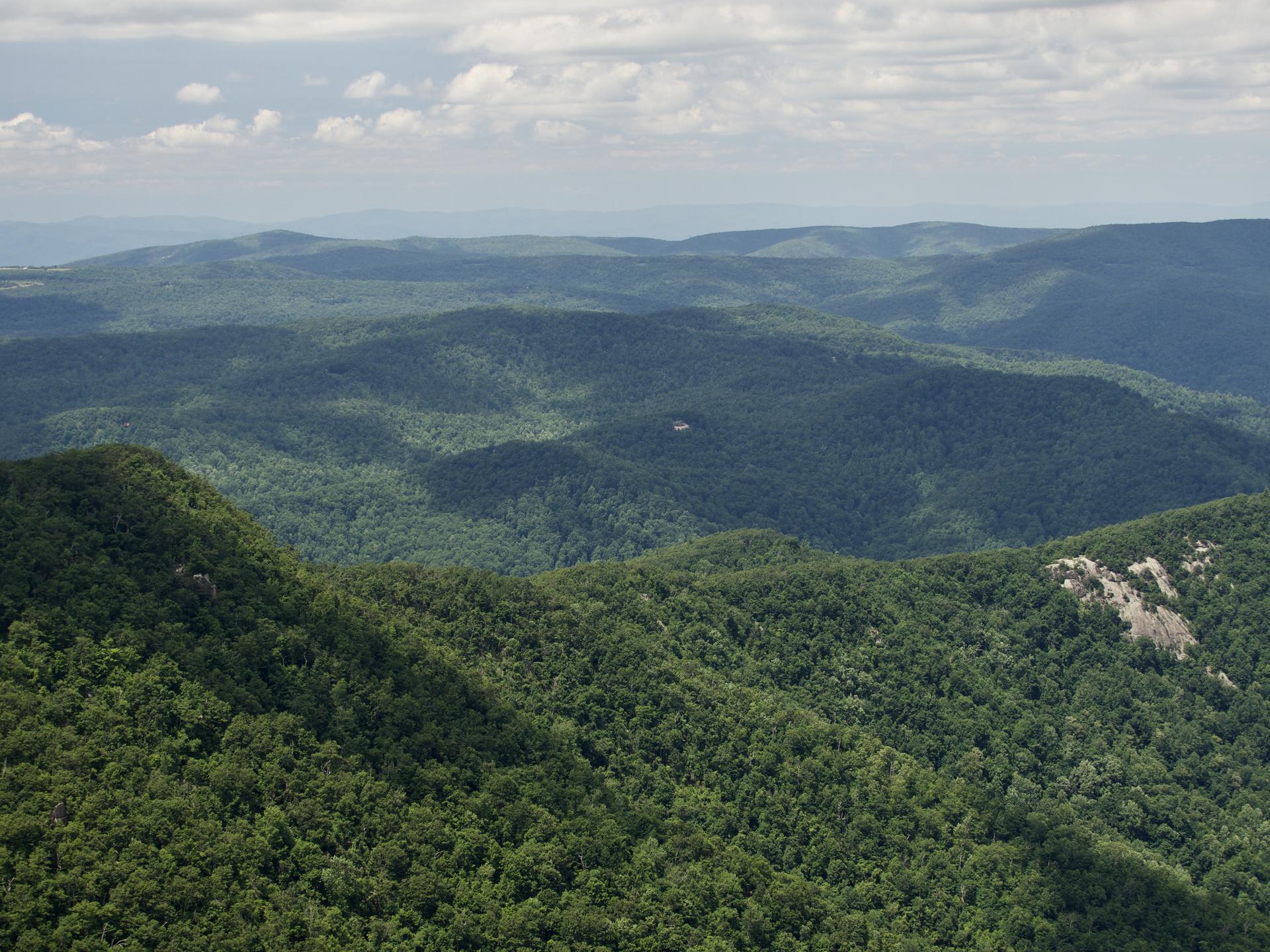 Vast green forested mountains and valleys stretch under a cloudy sky.