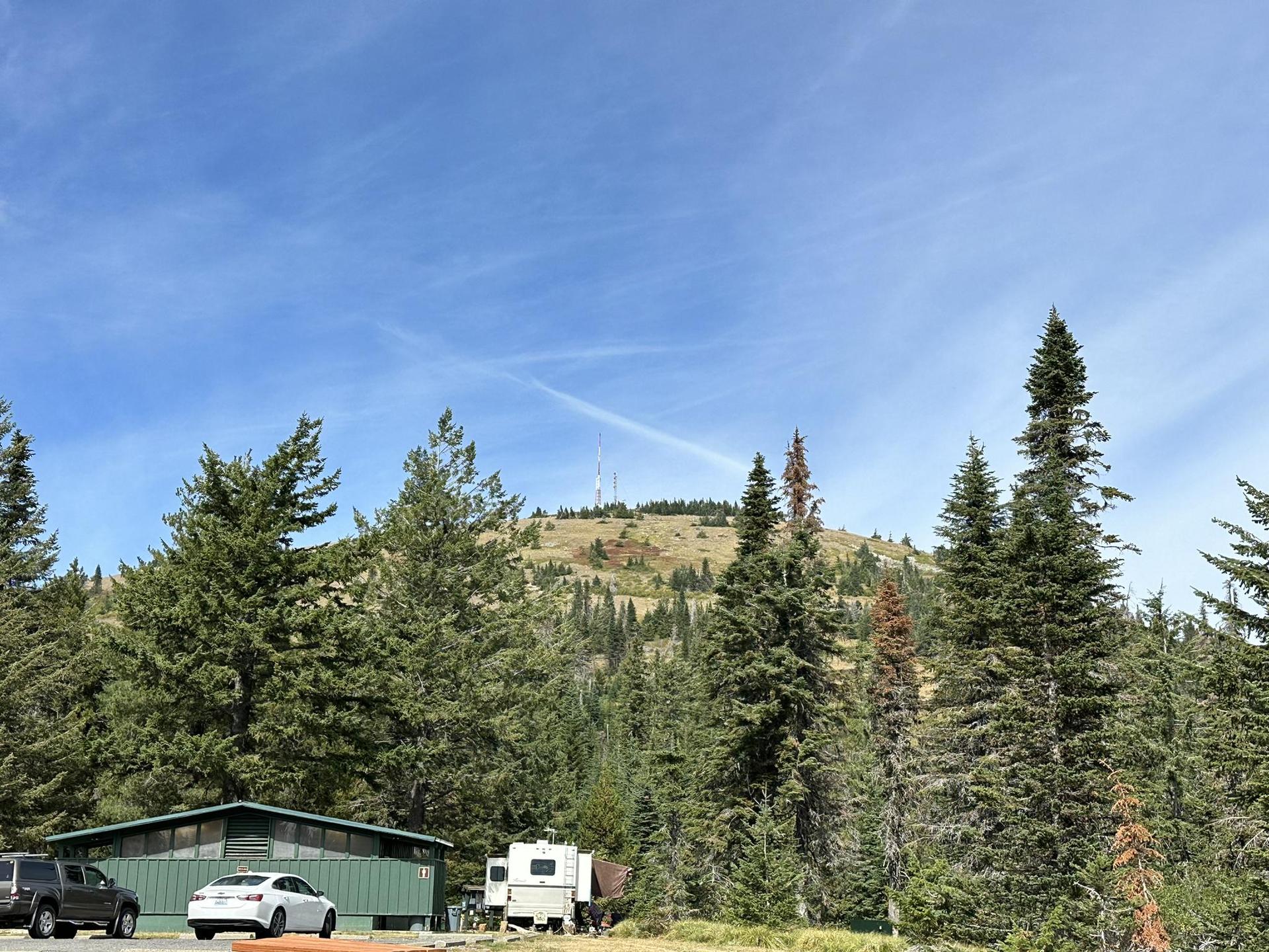 A mountain with communication towers overlooks a parking area and forest under a blue sky.
