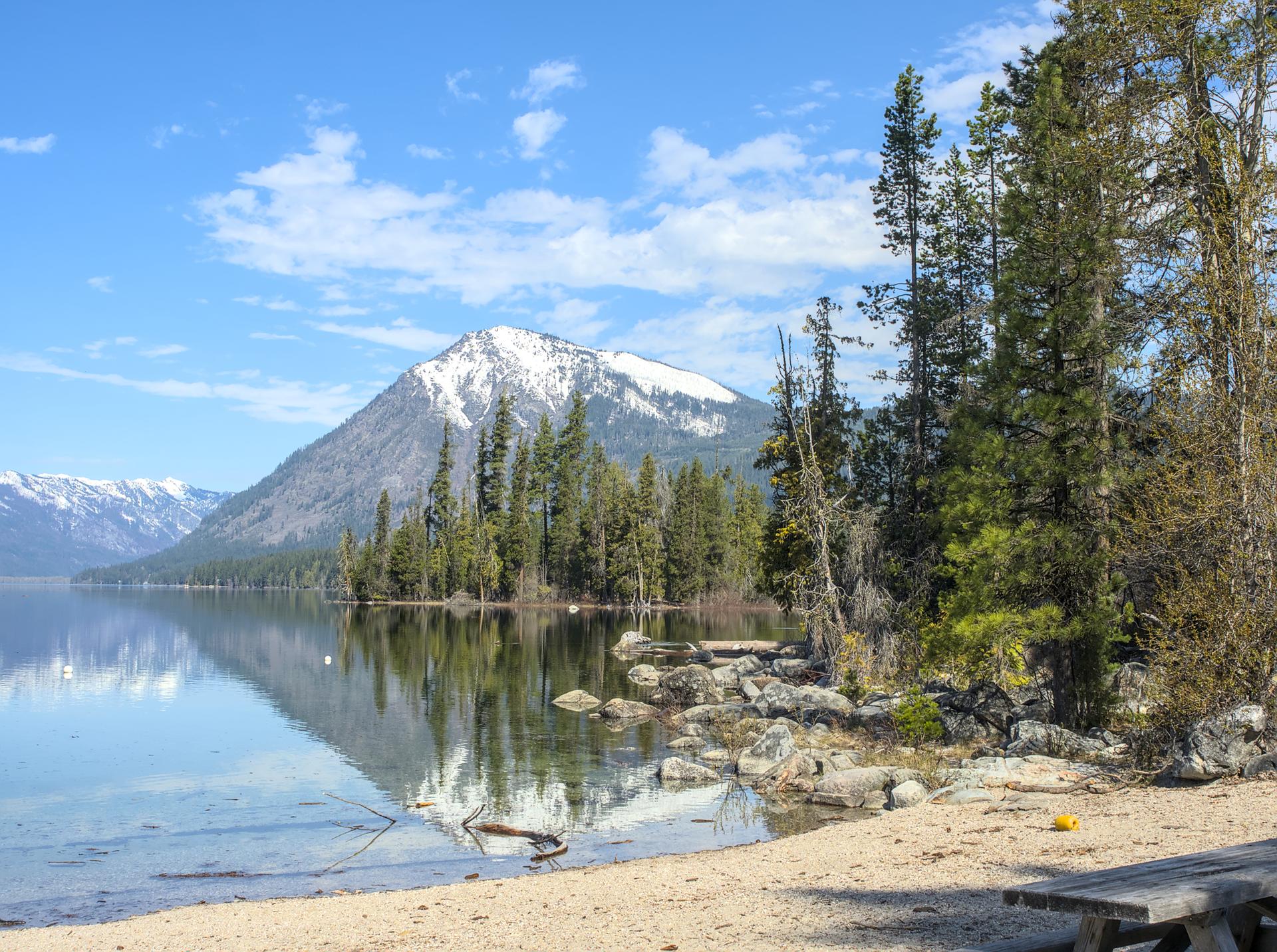 Sunny lake with snow-capped mountains, pine trees, and a sandy shore with a picnic table.