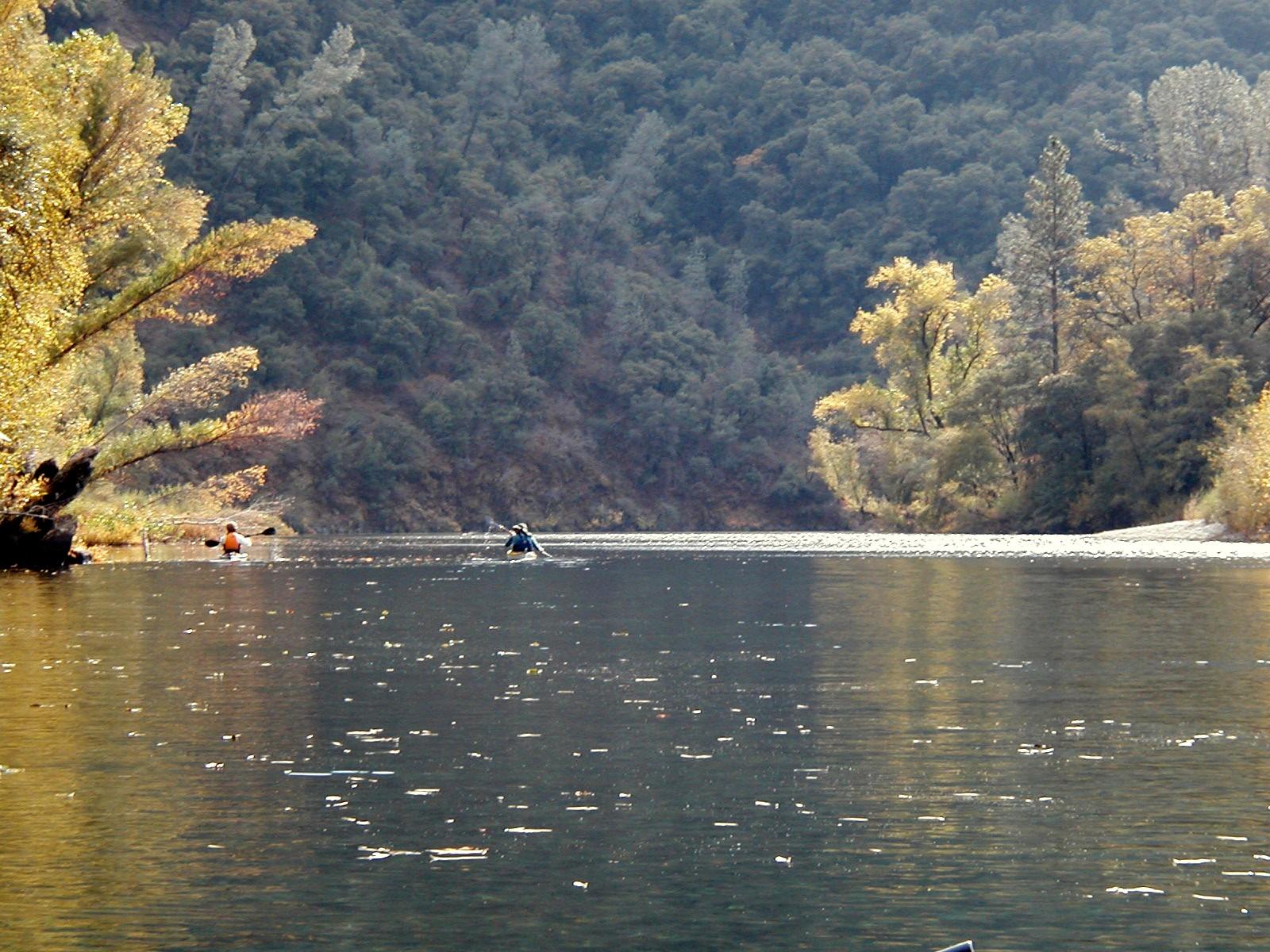 Two kayakers paddle on a river surrounded by autumn trees and forested hills.