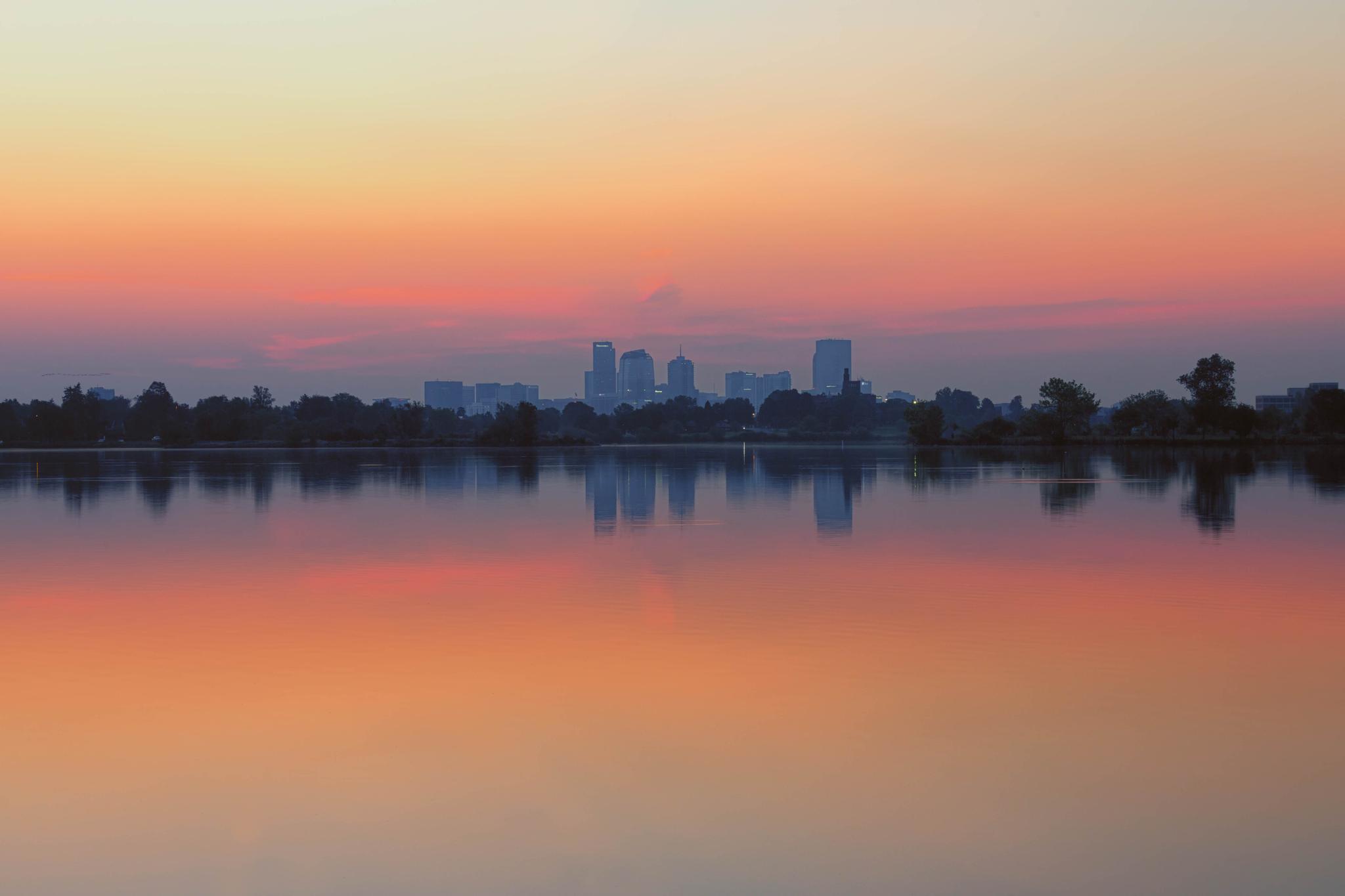 City skyline reflecting on a calm lake at sunset with orange and pink sky.